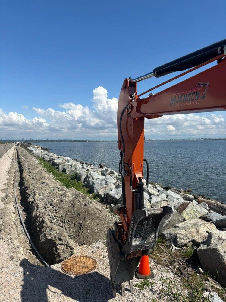 Excavator by water's edge on a construction site with clouds and shoreline in background.
