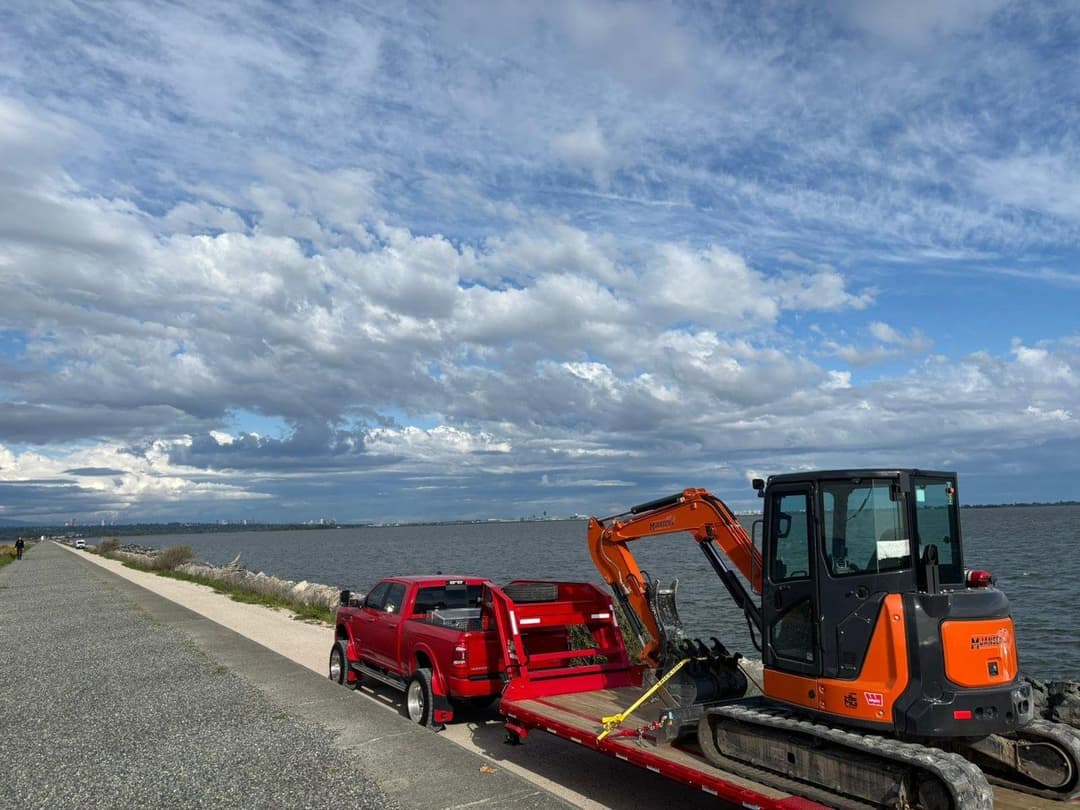 Excavator on trailer beside red pickup truck by waterfront under cloudy sky.