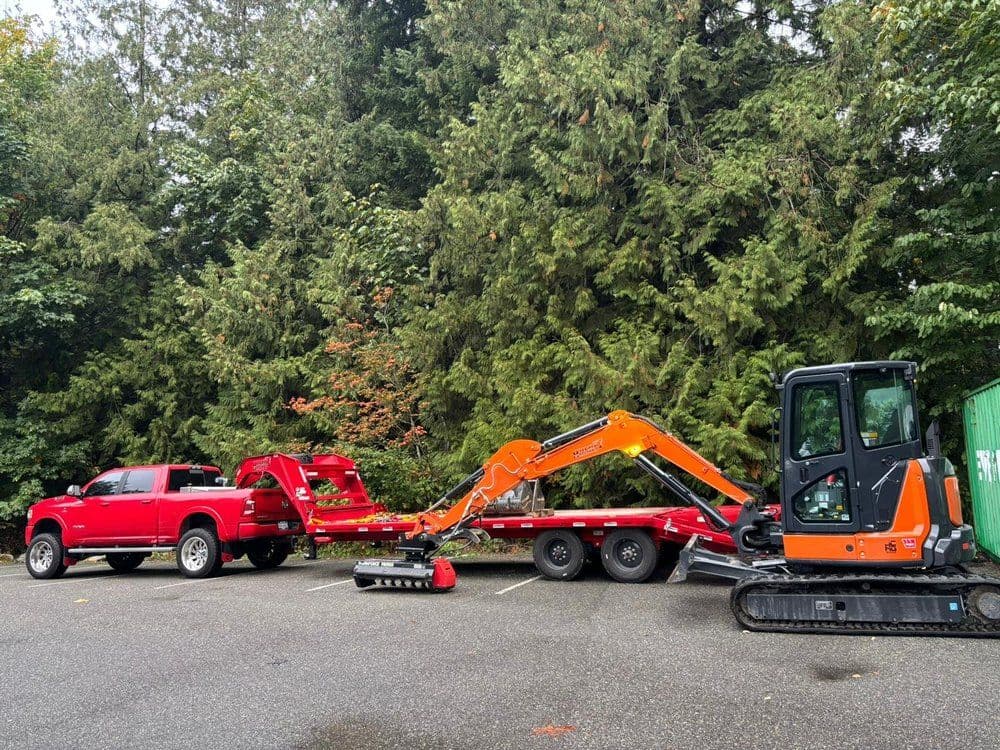 Red pickup truck towing excavation equipment in a forested area.