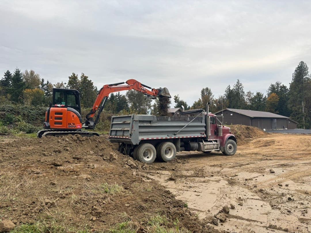 Excavator loading dirt into a truck at a construction site with trees in the background.