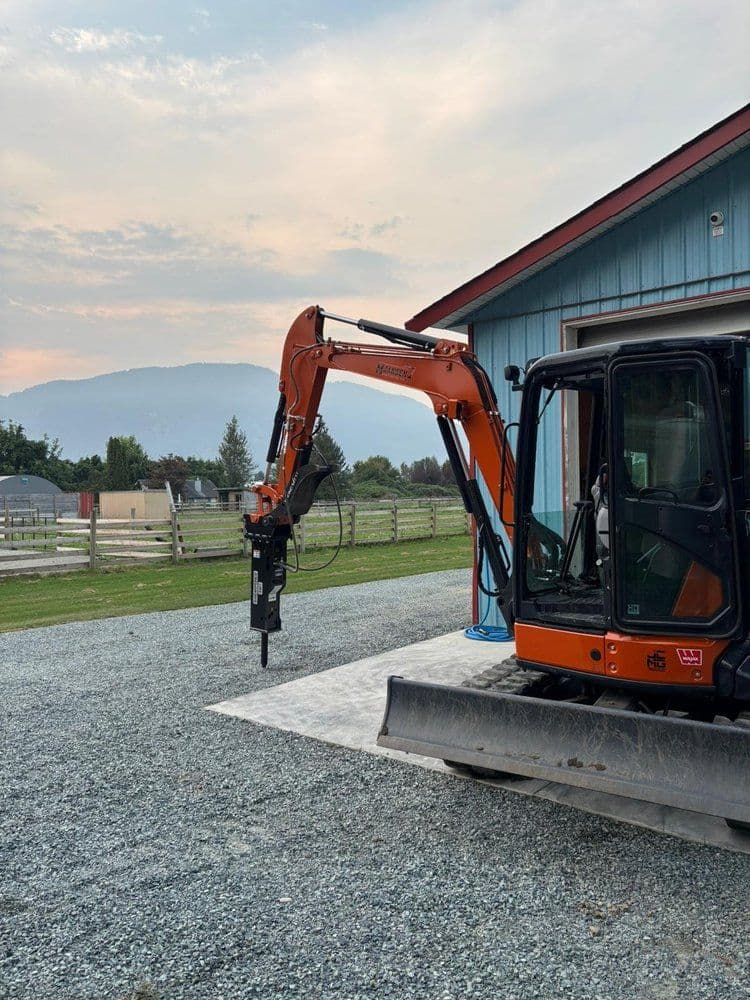 Excavator attachment outside a barn with mountains in the background, gravel surface.