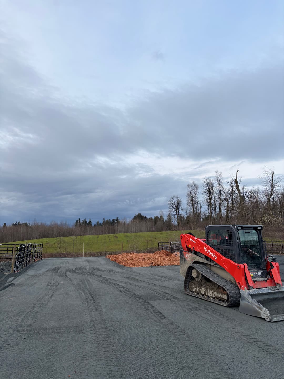 Kubota skid steer loader on gravel site with overcast sky and trees in the background.