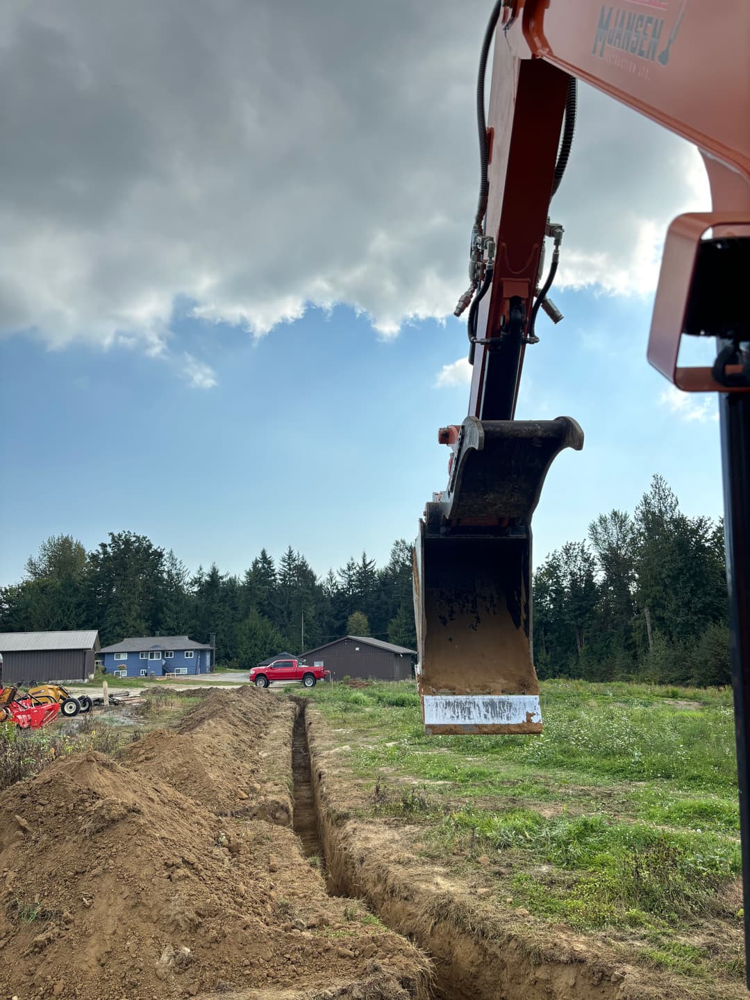 Excavator digging a trench on a rural site under a cloudy sky.