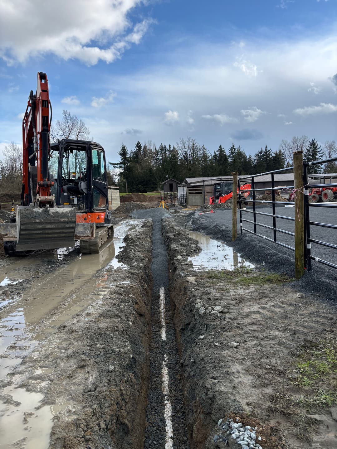 Excavator digging a trench on a construction site with cloudy sky and nearby equipment.
