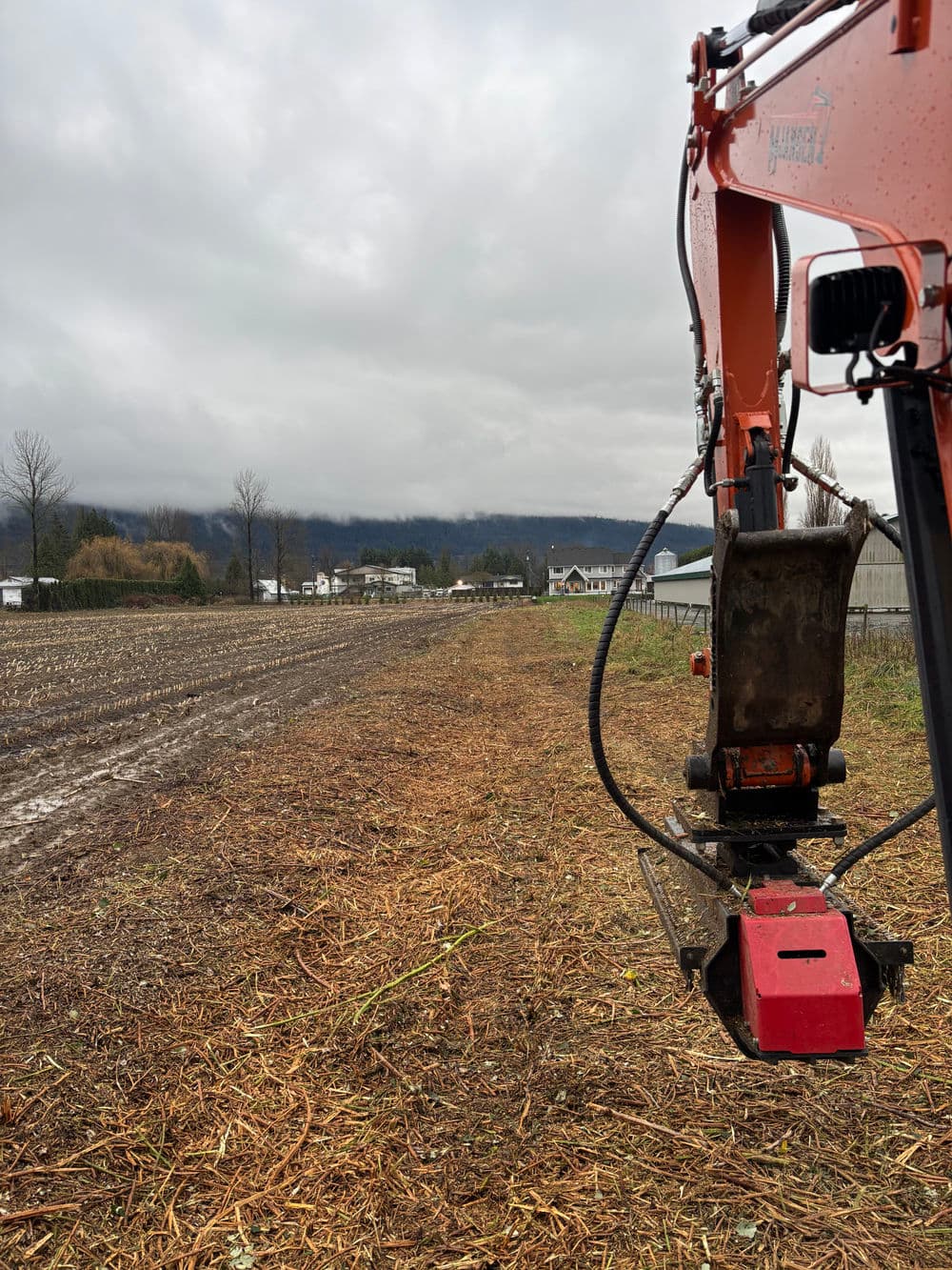 Excavator arm on a muddy field with cloudy sky and distant buildings in the background.