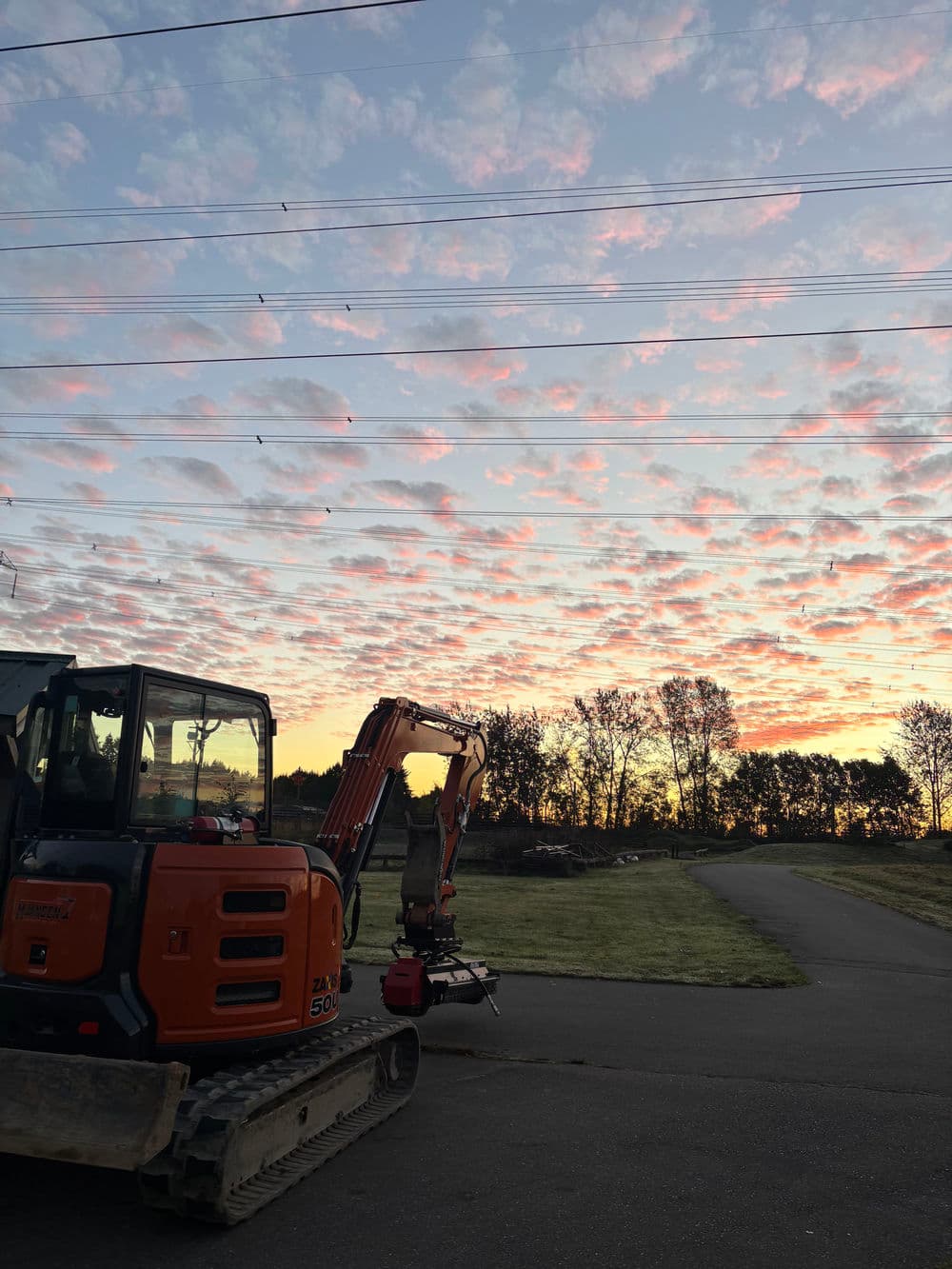 Excavator at sunset with colorful clouds and power lines in the background.