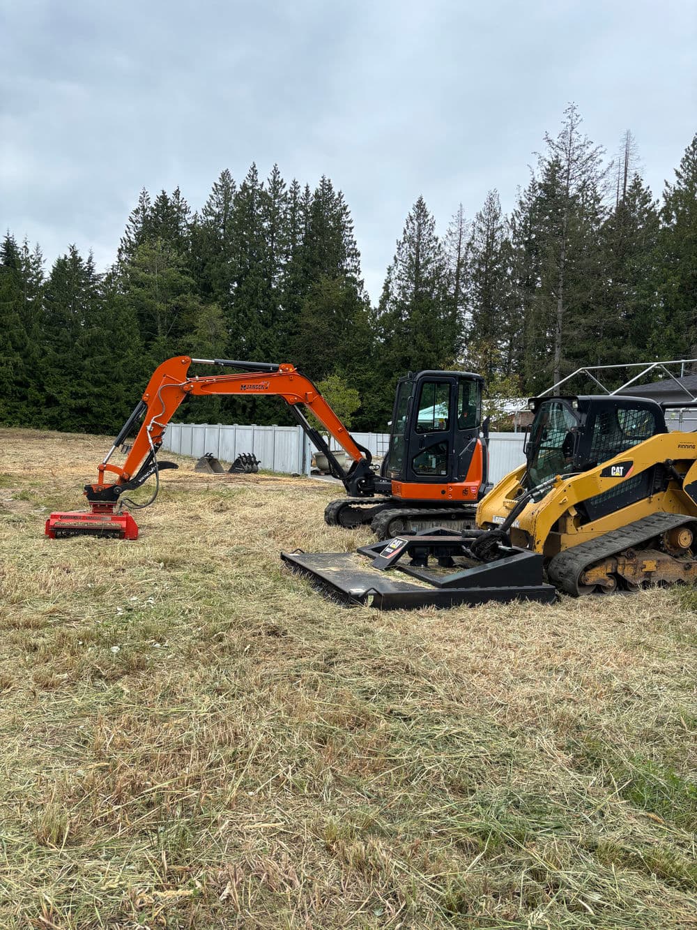 Excavators and skid steer on a clearing site surrounded by trees and grass.