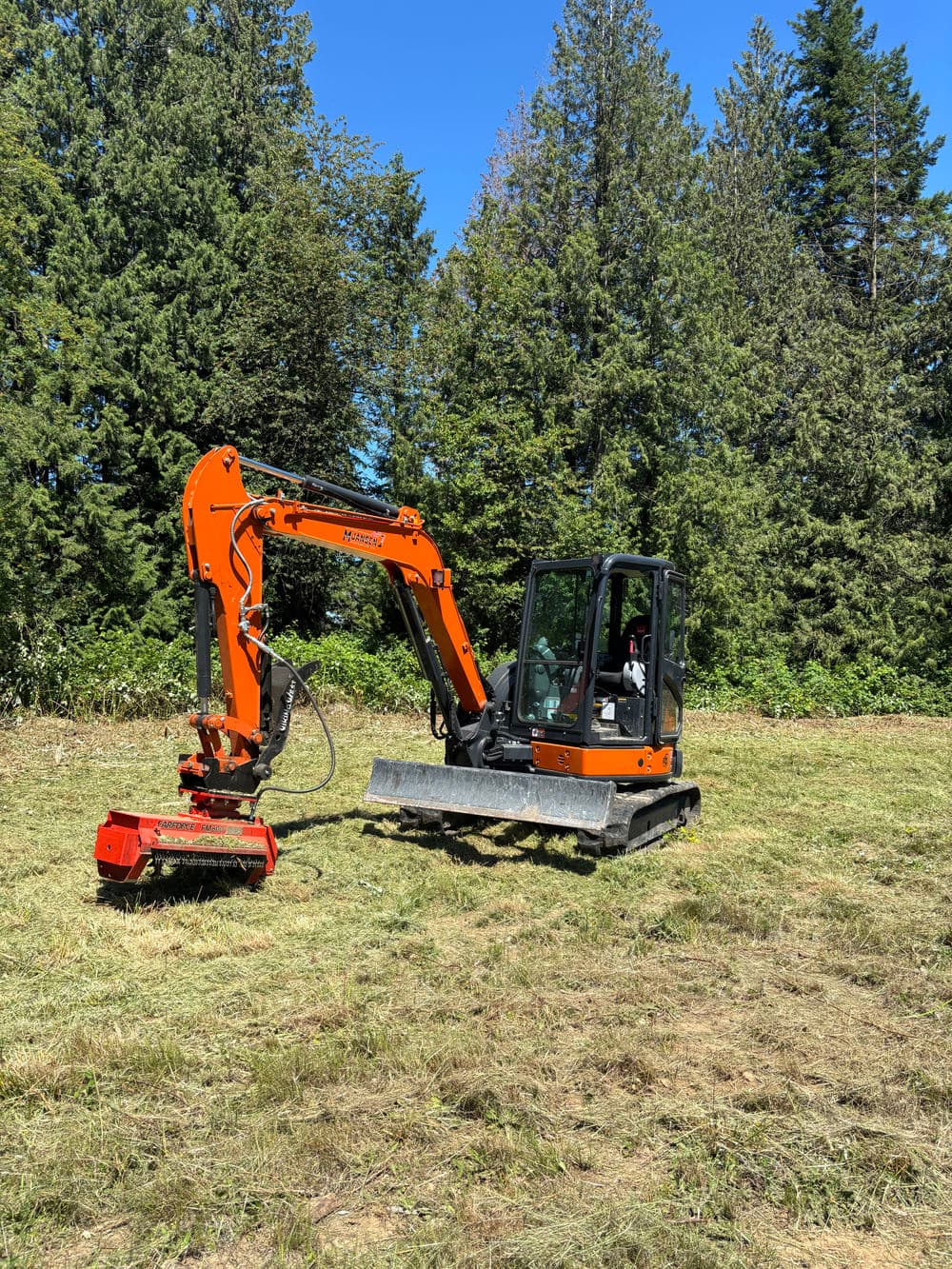 Excavator with brush cutter attachment operating in a green field surrounded by trees.