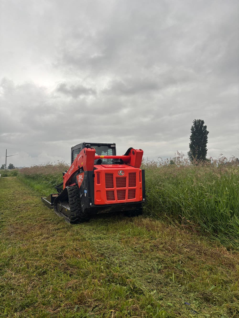 Red skid steer loader cutting grass in a field under cloudy skies.