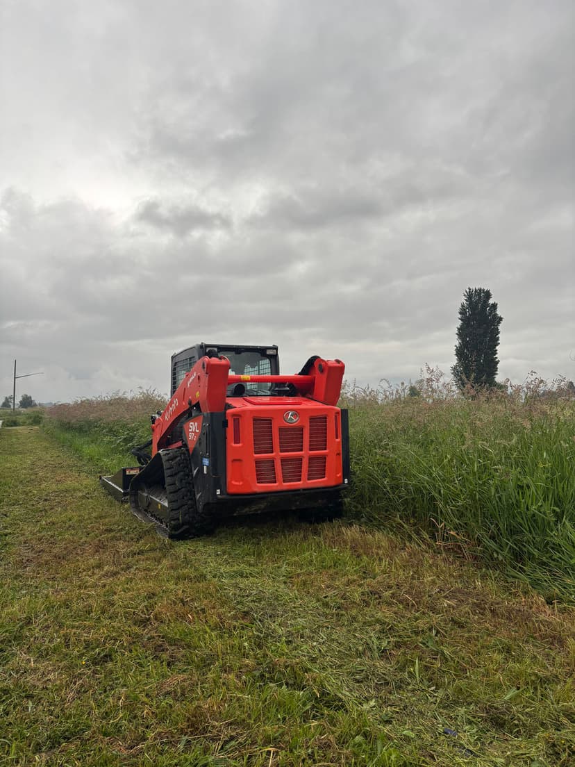 Red skid steer loader cutting grass in a field under a cloudy sky.